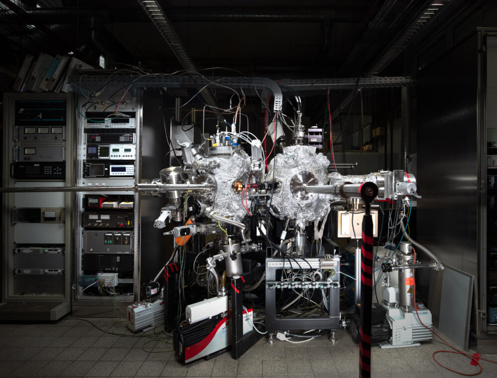A wide-angle, meticulously lit photograph of a complex Physical Vapour Deposition (PVD) system in a laboratory. The image shows the stainless steel vacuum chambers and magnetic slide valves, rendered with sharp detail and balanced lighting to highlight the engineering precision. Copyright Jan Söhlke.